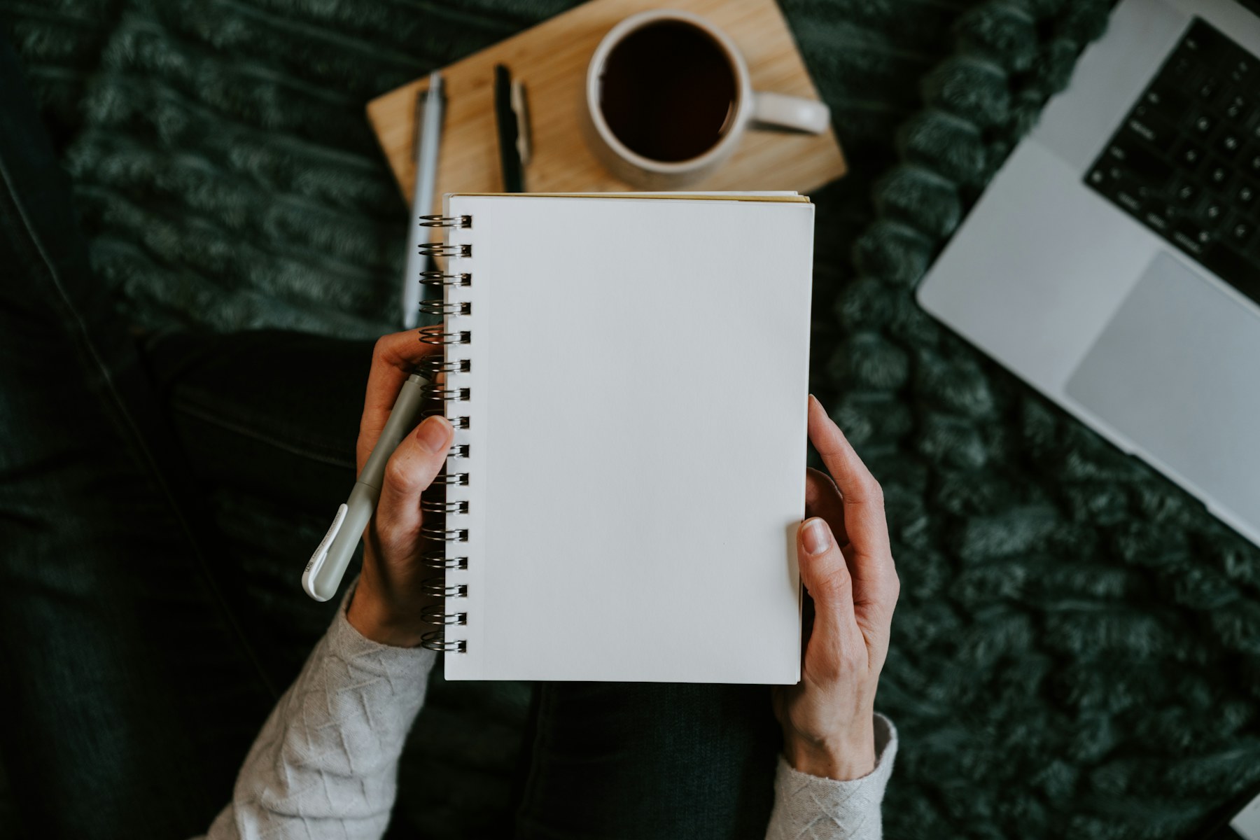A person sitting comfortably with a notebook, pen, coffee, and laptop